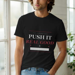 Puede incluir: Camiseta negra de cuello redondo con el texto "PUSH IT REAL GOOD" en blanco y rojo. La camiseta también presenta el texto "EST. COMMIT 2005" y "GIFT EDITION". La camiseta la lleva una persona.