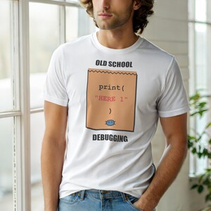 Pode incluir: Camiseta branca com um gráfico castanho que diz "OLD SCHOOL" e "DEBUGGING". O gráfico inclui um trecho de código: "print("HERE 1")". Uma pequena carinha azul está na parte inferior do design, um design humorístico.