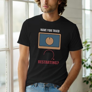 Op de afbeelding: Zwart T-shirt met de tekst "HAVE YOU TRIED RESTARTING?" boven een afbeelding van een computerscherm met een aan/uit-knop. Het scherm is blauw met een bruine rand en een bruine aan/uit-knop. Een rode gebogen pijl staat eronder.