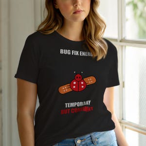 Op de afbeelding: Zwart T-shirt met een rood lieveheersbeestje ontwerp, een pleister en de tekst "BUG FIX ENERGY" en "TEMPORARY BUT CONFIDENT". Het lieveheersbeestje heeft witte stippen en staat in het midden van het shirt.