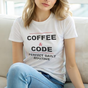 Op de afbeelding: Wit T-shirt met de tekst "COFFEE & CODE PERFECT DAILY ROUTINE" in zwart en rood. Het shirt heeft korte mouwen en wordt gedragen met een blauwe spijkerbroek. Het thema is vrijetijdskleding.