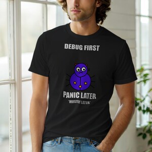 May include: Black t-shirt with the text "DEBUG FIRST PANIC LATER 'MOSTLY LATER'" and a purple cartoon bug with a worried expression. The shirt is a crew neck style.