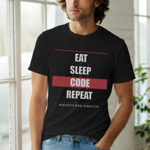 Op de afbeelding: Zwart T-shirt met de woorden "EAT SLEEP CODE REPEAT" in witte letters, met een rode balk onder het woord "CODE". Daaronder staat de zin "DEVELOPER MODE ALWAYS ON".