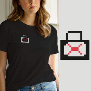 May include: A black short-sleeved t-shirt featuring a small pixel art graphic on the left chest. The graphic depicts a light gray rectangular bag with a black outline and a red pixelated cross symbol centered on the front of the bag design.