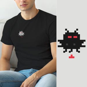 May include: A black short-sleeved t-shirt featuring a small pixel art graphic on the left chest. The graphic depicts a stylised creature with a black body, two red rectangular eyes, and a small red T-shaped element positioned below the figure.