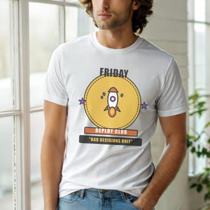 Op de afbeelding: Wit t-shirt met een afbeelding van een raket in een gele cirkel, het woord "FRIDAY" erboven en "DEPLOY CLUB" en "BAD DECISIONS ONLY" eronder. Het ontwerp bevat paarse sterren en oranje accenten.