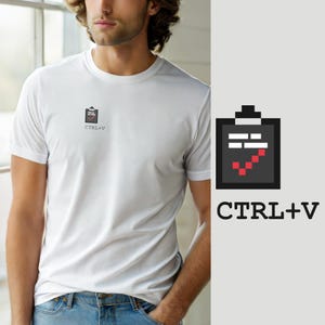 May include: A white short-sleeved t-shirt featuring a small graphic on the left chest. The graphic depicts a dark grey pixelated clipboard icon with white horizontal lines and a red pixelated checkmark, accompanied by the black text CTRL+V.