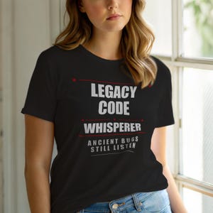 Könnte beinhalten: Schwarzes T-Shirt mit weißem Text: "LEGACY CODE WHISPERER" über "ANCIENT BUGS STILL LISTEN". Der Text ist fett und ohne Serifen. Rote Sternakzente befinden sich über dem Text. Das Shirt besteht aus weichem Material.
