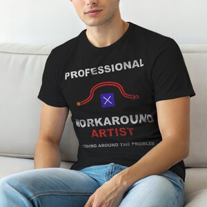 Op de afbeelding: Zwart t-shirt met de tekst "PROFESSIONAL WORKAROUND ARTIST" in wit en rood. Het shirt heeft een grafisch ontwerp met een rode lijn en een blauw vierkant. De zin "CODING AROUND THE PROBLEM" staat eronder gedrukt.