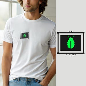 Può includere: Una t-shirt bianca a maniche corte con una piccola grafica rettangolare sul petto. La grafica raffigura una cornice nera contenente l'icona di uno scarabeo verde brillante. Sotto la cornice, il testo recita > FIXED in un semplice font nero.