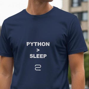 Op de afbeelding: Marineblauw T-shirt met witte tekst "PYTHON > SLEEP" en het nummer 2 eronder. Het shirt met korte mouwen is gemaakt van een zacht materiaal. De achtergrond toont een vage stedelijke omgeving.