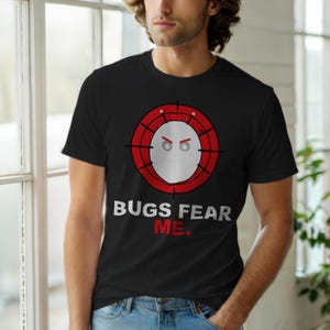 May include: Black crew neck t-shirt with the text "BUGS FEAR ME." The shirt features a graphic of a white face with red eyes and a red target design around it. The design is centered on the front of the shirt.