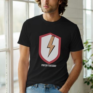 Op de afbeelding: Zwart T-shirt met ronde hals en een schildontwerp met een bruine en witte bliksem. De tekst "PATCH TUESDAY" staat eronder gedrukt. Het T-shirt is gemaakt van zacht materiaal.