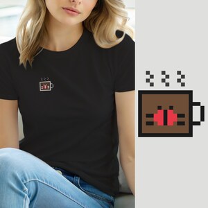 Könnte beinhalten: Ein schwarzes T-Shirt mit kurzen Ärmeln und einer kleinen Pixel-Art-Grafik auf der linken Brust. Die Grafik zeigt eine braune, quadratische Tasse mit schwarzem Henkel, die ein rot-schwarzes Marienkäfer-Design enthält, mit drei kleinen Pixel-Dampfwolken.