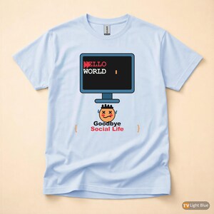 Op de afbeelding: Lichtblauw T-shirt met een computerscherm afbeelding met "HELLO WORLD" in rode en witte tekst. Onder het scherm staat een cartoongezicht en de tekst "Goodbye Social Life". Het shirt is van het merk Gildan.