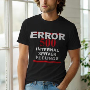 May include: Black t-shirt with the text "ERROR 500 INTERNAL SERVER FEELINGS" in white and red. The text is centered on the front of the shirt. The shirt is being worn by a person.