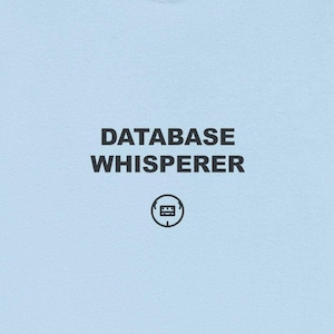 May include: Pale blue t-shirt featuring the text "DATABASE WHISPERER" in black, with a small circular graphic beneath. The shirt is made of a soft fabric and has short sleeves.