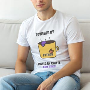 Puede incluir: Camiseta blanca con el texto "POWERED BY PYTHON" sobre un gráfico de una taza de café amarilla. La taza tiene un bicho morado y la palabra "PYTHON". Debajo está el texto "FUELED BY COFFEE AND BUGS".