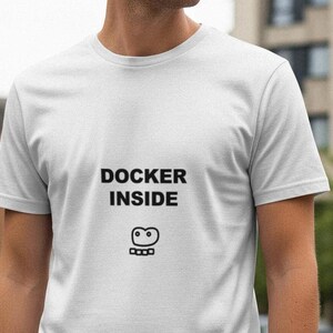 May include: White t-shirt with the text "DOCKER INSIDE" in black block letters above a small graphic. The shirt is short-sleeved and appears to be made of a soft material. The person wearing the shirt is standing outside.