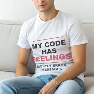 Op de afbeelding: Wit T-shirt met de tekst "MY CODE HAS FEELINGS MOSTLY ERROR MESSAGES" in zwart en rood. Het shirt heeft een ronde hals.