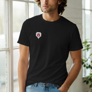 May include: Black t-shirt with a small, pixelated red and white graphic on the chest. The graphic appears to be a stylized shield or emblem. The shirt is made of a soft material and is being worn by a person.