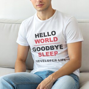 Op de afbeelding: Wit T-shirt met de tekst "HELLO WORLD GOODBYE SLEEP DEVELOPER LIFE" in zwart en rood. Het shirt heeft korte mouwen en wordt gedragen door een persoon die een blauwe spijkerbroek draagt.