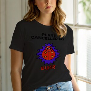May include: Black t-shirt with the text "PLANS CANCELLED BY BUGS." The shirt features a cartoon ladybug design in red, orange, and yellow, surrounded by a purple burst.