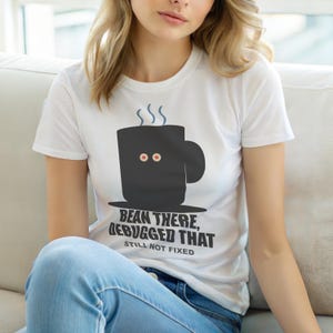 Puede incluir: Camiseta blanca con un diseño de taza de café negra, ojos rojos y vapor. El texto "BEAN THERE, DEBUGGED THAT, STILL NOT FIXED" está impreso debajo. La camiseta es de manga corta.