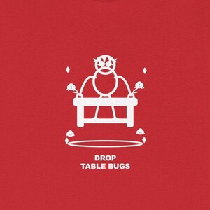 May include: A red t-shirt with a white graphic design featuring a stylized figure above a table, surrounded by diamond shapes and a circular base. The text "DROP TABLE BUGS" is printed below the table.