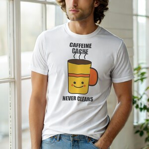 Op de afbeelding: Wit t-shirt met een cartoon koffiekopje. De gele beker heeft een lachend gezicht en de tekst "CAFFEINE CACHE NEVER CLEARS" in het zwart. De beker heeft een bruine bovenkant en een oranje handvat.