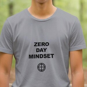 May include: A gray crew neck t-shirt with the words "ZERO DAY MINDSET" printed in black, along with a target symbol. The shirt is made of a soft material and is suitable for casual wear.