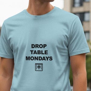 Può includere: Una maglietta azzurra con la scritta nera "DROP TABLE MONDAYS" e una piccola grafica. La maglietta è realizzata in materiale morbido e ha maniche corte. La persona è in piedi fuori da un edificio.