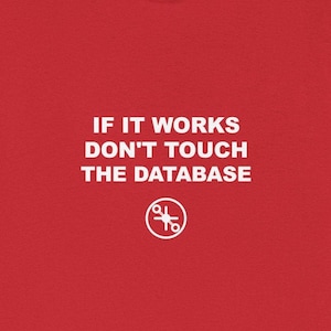 May include: A red t-shirt with white text that reads "IF IT WORKS DON'T TOUCH THE DATABASE". Below the text is a white circular graphic with a scissors design. The t-shirt is made of a soft material.