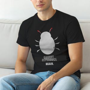 Op de afbeelding: Zwart t-shirt met een witte insectenafbeelding met rode antennes en de tekst "CANNOT REPRODUCE AGAIN." Het ontwerp is gecentreerd. Het t-shirt is gemaakt van een zacht materiaal.