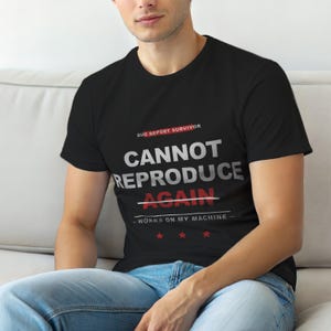 Op de afbeelding: Zwart t-shirt met witte en rode tekst. De tekst luidt "BUG REPORT SURVIVOR CANNOT REPRODUCE AGAIN - WORKS ON MY MACHINE" met drie rode sterren eronder. Het shirt heeft korte mouwen.