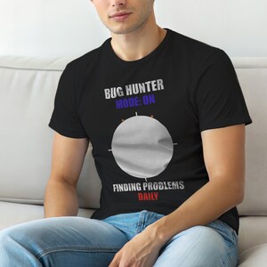 May include: Black t-shirt featuring the text "BUG HUNTER MODE: ON" above a grey circle with crosshairs. Below the circle, the text reads "FINDING PROBLEMS DAILY". The shirt is worn by a person.