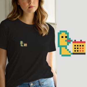 Pode incluir: Uma t-shirt preta de manga curta com um pequeno gráfico de pixel art no peito. O gráfico mostra uma personagem amarela e azul-petróleo com uma boca vermelha posicionada ao lado de um calendário amarelo com detalhes de grelha pretos e vermelhos.