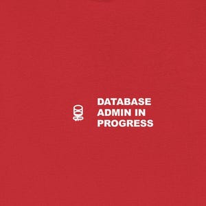 Può includere: Una maglietta rossa con la scritta bianca "DATABASE ADMIN IN PROGRESS". La maglietta presenta anche una piccola grafica a sinistra del testo. La maglietta è realizzata in un materiale morbido.