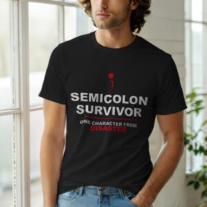 Op de afbeelding: Zwart T-shirt met ronde hals en de witte tekst "SEMICOLON SURVIVOR", een rood puntkomma-symbool en de rode woorden "ONE CHARACTER FROM DISASTER". Het shirt is gemaakt van een zacht materiaal.