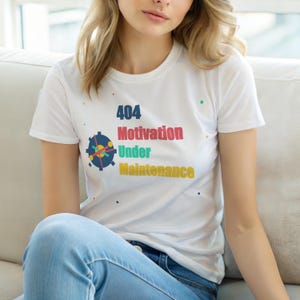 Peut inclure: T-shirt blanc avec l'inscription "404 Motivation Under Maintenance" en plusieurs couleurs. Le design comprend un engrenage et des points colorés. Le t-shirt est porté avec un jean bleu.