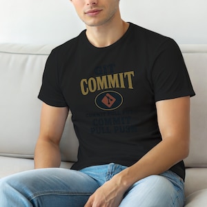 Op de afbeelding: Een zwart t-shirt met korte mouwen voorzien van de tekst GIT COMMIT in goudkleurige letters, een centraal oranje Git-logo in een ovaal, en de herhaalde tekst COMMIT PULL PUSH in donkerblauwe letters op de voorzijde.