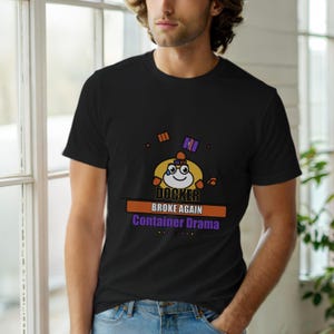 以下が含まれることがあります： 黒いTシャツ。漫画のキャラクターと「DOCKER BROKE AGAIN Container Drama」の文字がプリントされています。デザインはオレンジ、紫、黄色を使用。ユーモラスな技術テーマです。