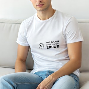 May include: White t-shirt with the text "404 BRAIN INITIALIZATION ERROR" in black. The shirt features a smiling face graphic. The person is wearing blue jeans.