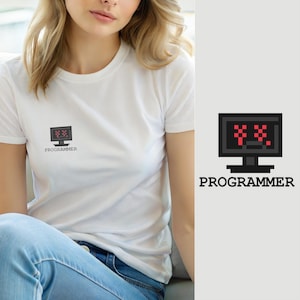 Op de afbeelding: Een wit T-shirt met korte mouwen en een kleine afbeelding op de borst van een zwarte computermonitor met een gepixeld rood scherm met twee X-vormige ogen en het woord PROGRAMMER in zwarte hoofdletters eronder gedrukt.