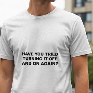 May include: White t-shirt with the text "HAVE YOU TRIED TURNING IT OFF AND ON AGAIN?" in black. The shirt has short sleeves and a crew neck. The person wearing the shirt is standing outside in front of a building.