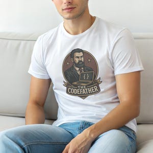 Peut inclure: Un t-shirt blanc à manches courtes arborant un motif circulaire représentant un homme barbu en costume derrière un ordinateur portable affichant des accolades. Sous l'homme, une bannière affiche le texte THE CODEFATHER en lettres capitales.