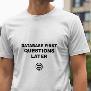 May include: White t-shirt with the text "DATABASE FIRST QUESTIONS LATER" in black, along with a circular graphic. The shirt is short-sleeved and appears to be made of a soft material. The person is standing outside.