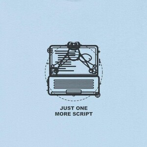 Op de afbeelding: Lichtblauw T-shirt met een zwarte afbeelding van een laptop en een pen. De tekst "JUST ONE MORE SCRIPT" staat eronder gedrukt. Het T-shirt is gemaakt van een zacht materiaal.