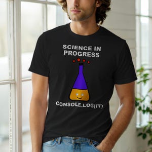 Op de afbeelding: Zwart T-shirt met de tekst "SCIENCE IN PROGRESS" boven een cartoon beker met een paarse bovenkant en gele basis. De tekst "CONSOLE.LOG(IT)" staat onder de beker.
