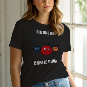 Op de afbeelding: Zwart T-shirt met de tekst "FIX ONE BUG CREATE SEVEN". Het shirt heeft een afbeelding van drie cartoon lieveheersbeestjes in rood, blauw en oranje. Het lieveheersbeestje in het midden is rood met zwarte stippen.